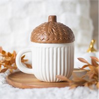 White mug with a brown acorn lid on a wooden coaster, surrounded by autumn leaves.