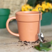 Terracotta mug with a spoon on a wooden surface with soil and plants in the background